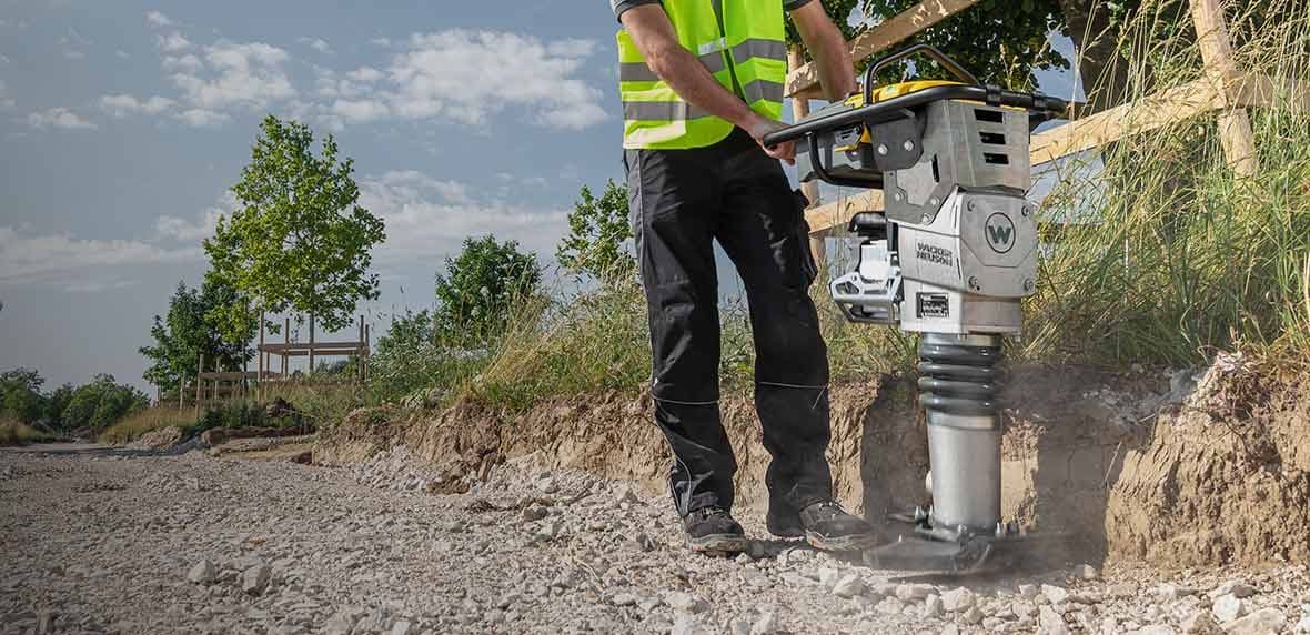 Werknemer met veiligheidsvest bedient een Wacker Neuson stamper op een stenige bouwstrook.