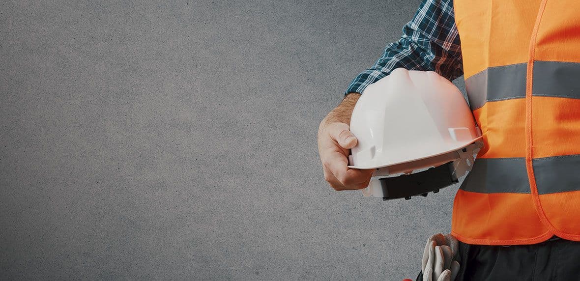 Bouwmedewerker met oranje veiligheidsvest houdt een witte bouwhelm tegen een grijze achtergrond.