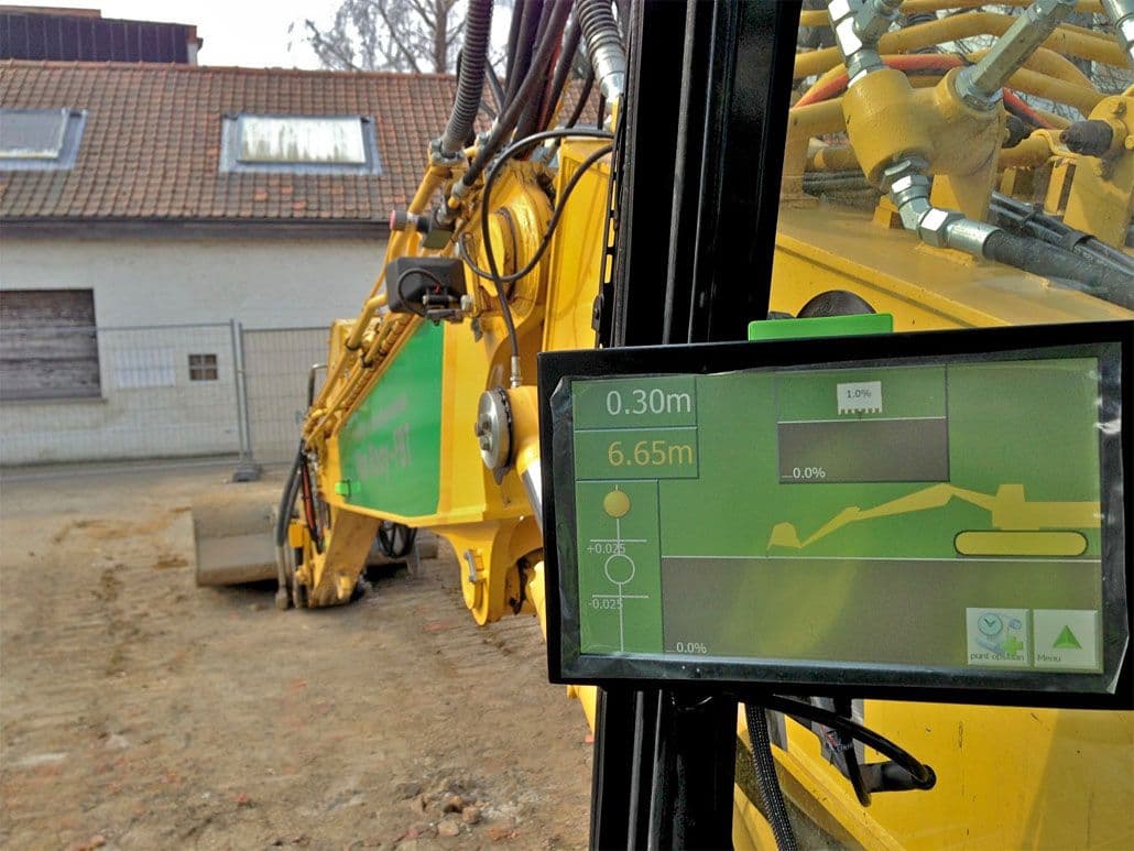 Bediening van een DigPilot 3D-graafmachinebesturingssysteem met zicht op het scherm in de cabine en een geel hydraulisch graafarm buiten.