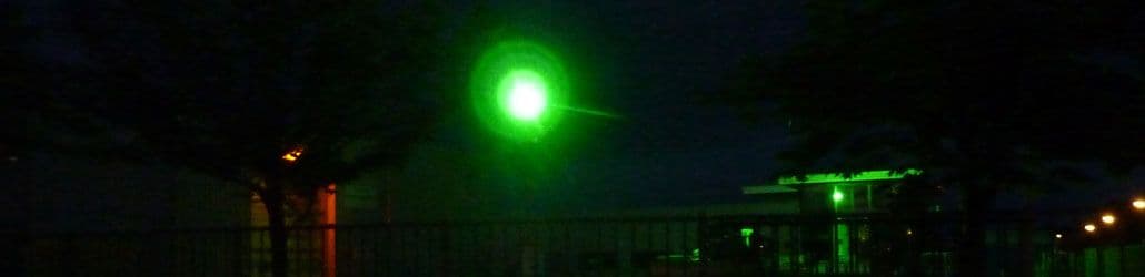 Groene LED-bouwlamp verlicht een industrieterrein in het donker, met silhouetten van bomen en gebouwen op de achtergrond.