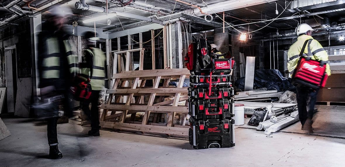 Drie personen met veiligheidsvesten lopen in bouwruimte met Milwaukee gereedschapskoffers.