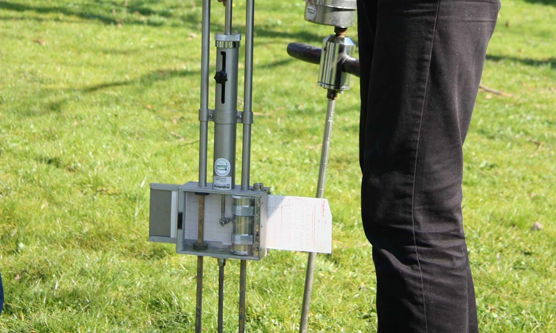 Persoon voert een bodemonderzoek uit met een handmatige penetrograaf op een grasveld met herfstbladeren.