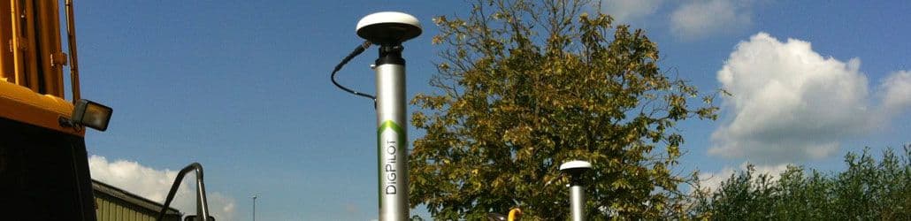 Twee DigPilot GPS-antennes op metalen palen bij een graafmachine, met een boom en blauwe lucht op de achtergrond.
