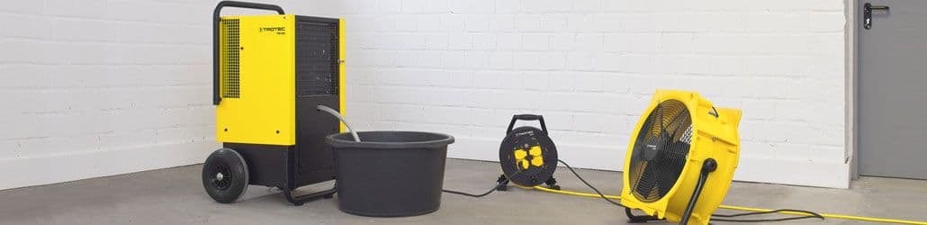 Opstelling van een bouwdroger, ventilator en kabelhaspel in een lege ruimte, met een opvangbak voor condenswater aangesloten op de droger.