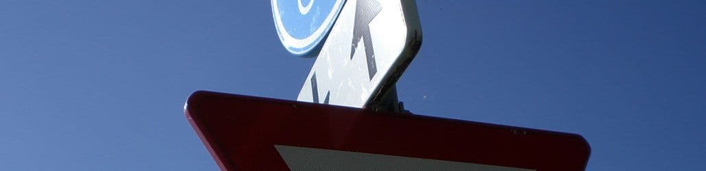 Laag camerastandpunt op twee verkeersborden tegen een blauwe lucht, met zicht op een omgekeerde driehoek (voorrang verlenen) en een gedeeltelijk zichtbaar blauw bord met pijl omhoog.
