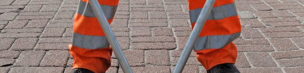 Close-up van de onderbenen van een persoon in een oranje reflectiebroek met zilverkleurige striping. De persoon staat op een straat met rode klinkers en houdt een metalen voorwerp vast.