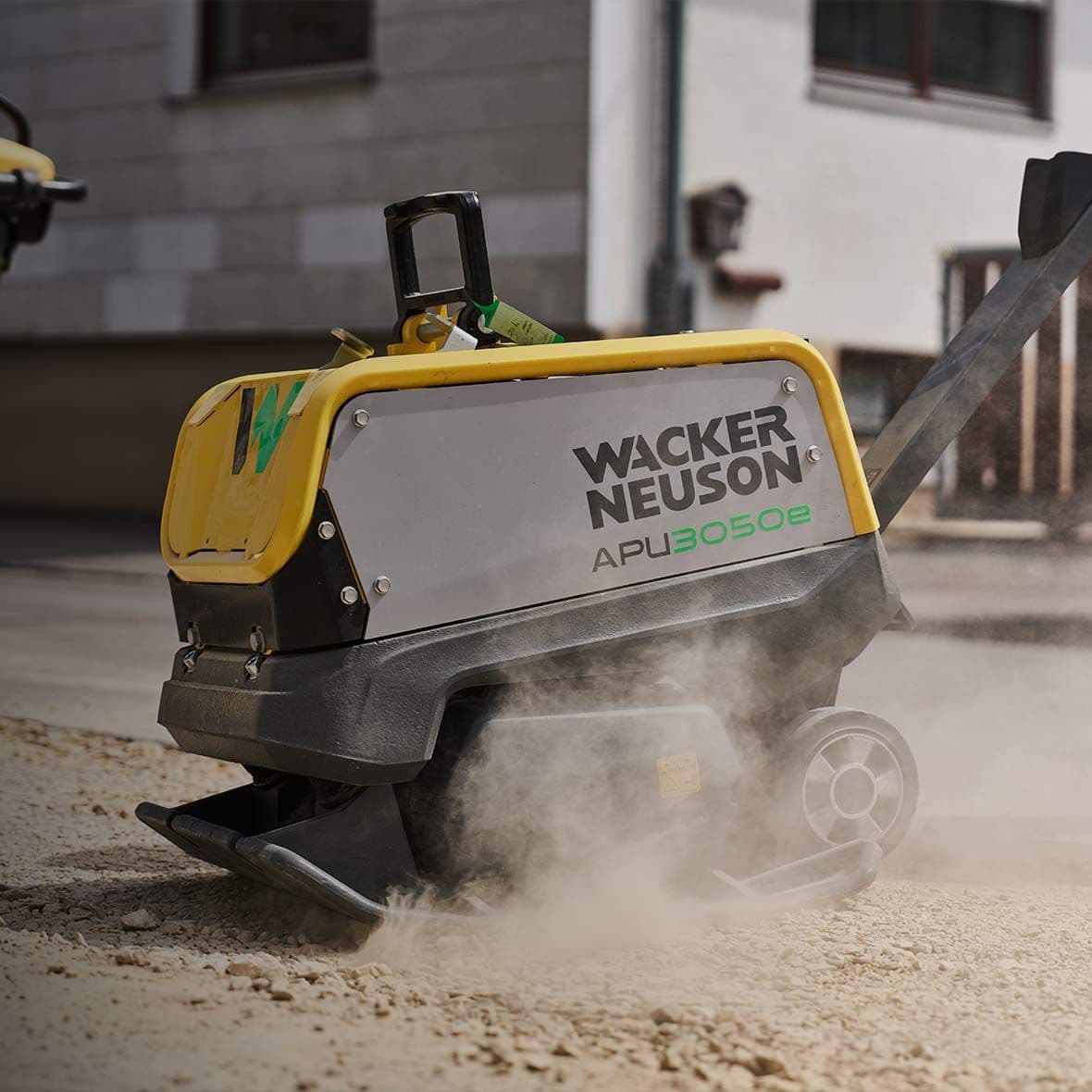 Gele en grijze Wacker Neuson APU3050e trilplaat die stof opwerpt op een bouwondergrond.