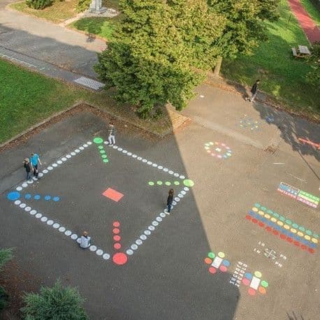 Schoolplein met kleurrijke speelvakken en spelpatronen op de grond, aangebracht met markeringsverf. Kinderen spelen op een groot 'Mens erger je niet'-spel.