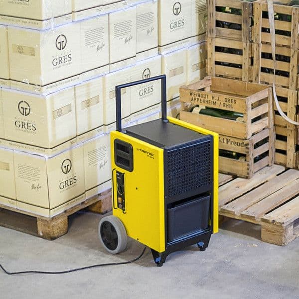 Gele Trotec bouwdroger geplaatst in een magazijn, tussen gestapelde wijnverpakkingen en houten pallets.