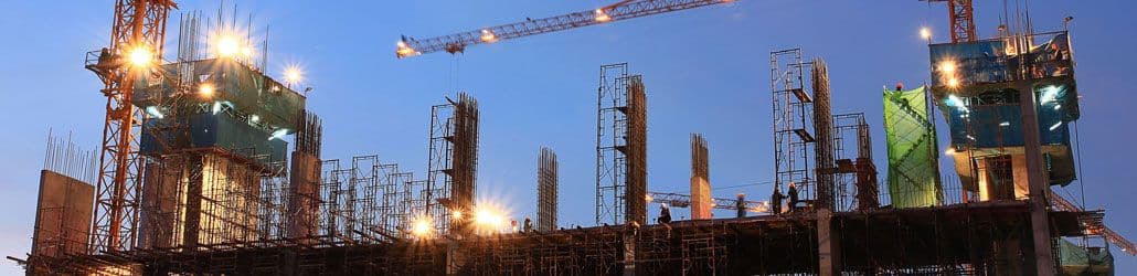 Bouwplaats bij schemering met steigers, hijskranen en meerdere heldere LED-bouwlampen die het terrein verlichten.