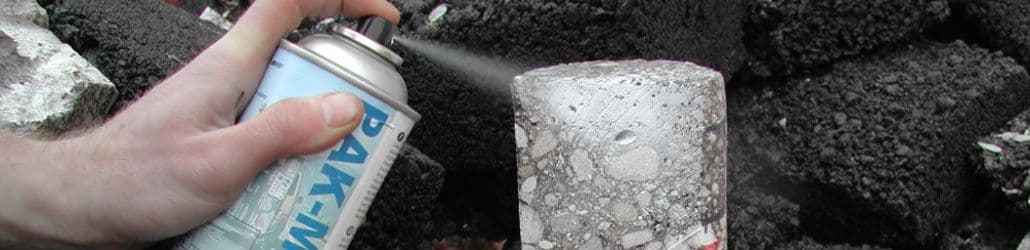 Hand spuit met een PAK-marker op een boorkern van asfalt of beton, met donkere asfaltblokken op de achtergrond.