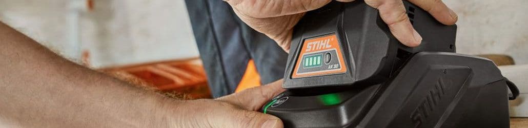 Close-up van een STIHL AK 30 accu die in een acculader wordt geplaatst, met zicht op de laadindicator en de handen van de gebruiker.