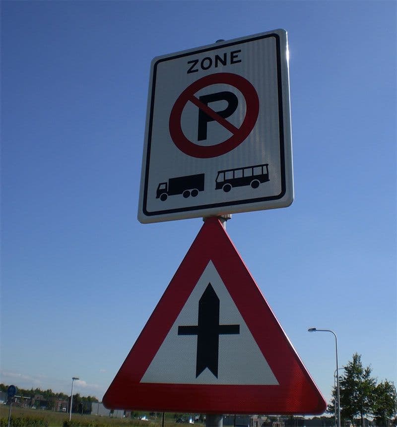 Verkeersbord met parkeerverbod voor vrachtwagens en bussen binnen een zone, geplaatst boven een waarschuwingsbord voor een kruising met zijwegen aan beide zijden.