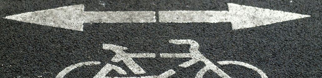 Wegmarkering van een fietssymbool met twee witte pijlen die tegengestelde richtingen aanduiden op een zwart asfalt.