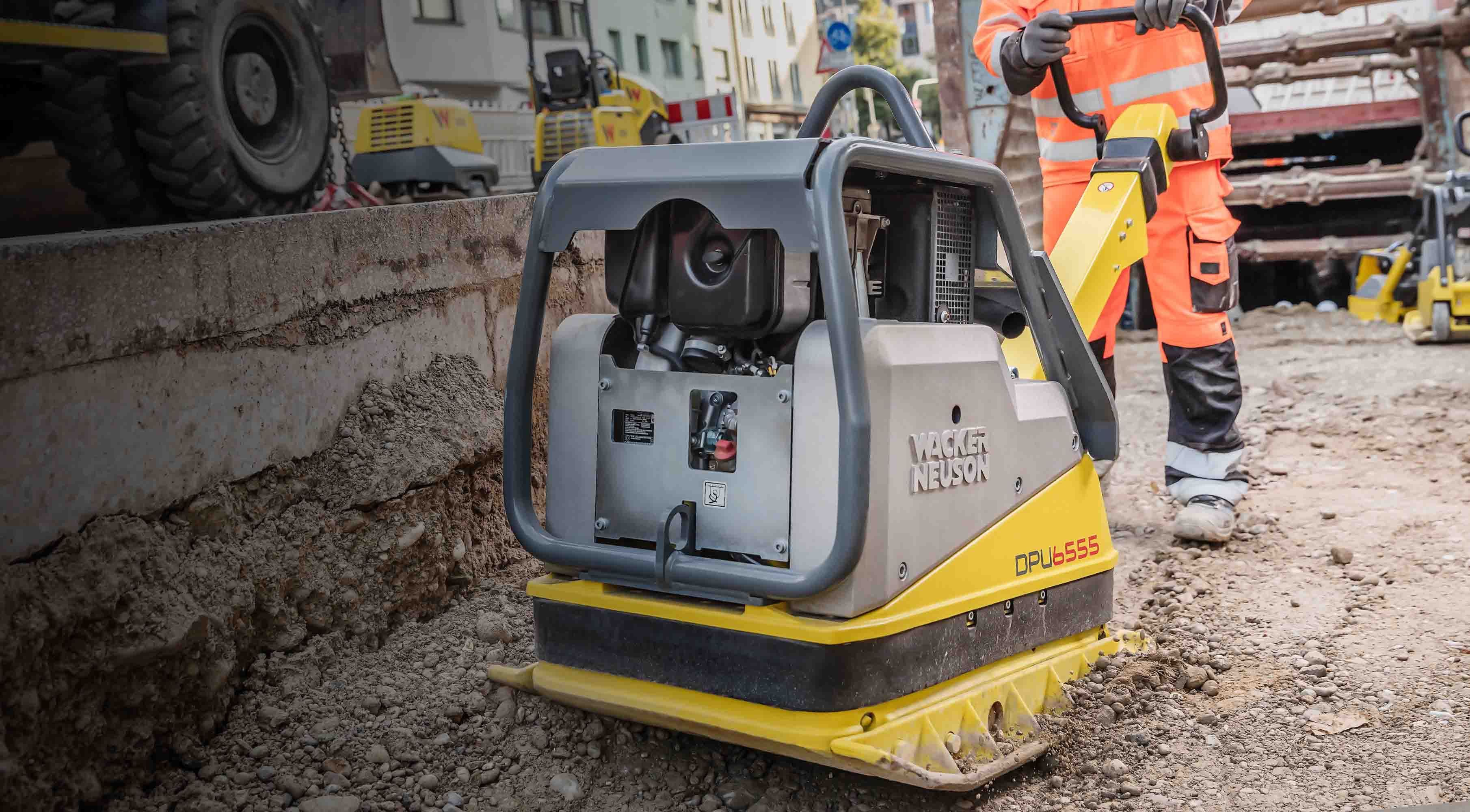 Alt-Tekst Generator zei: Bouwmedewerker bedient een gele Wacker Neuson trilplaat op een stenige ondergrond.