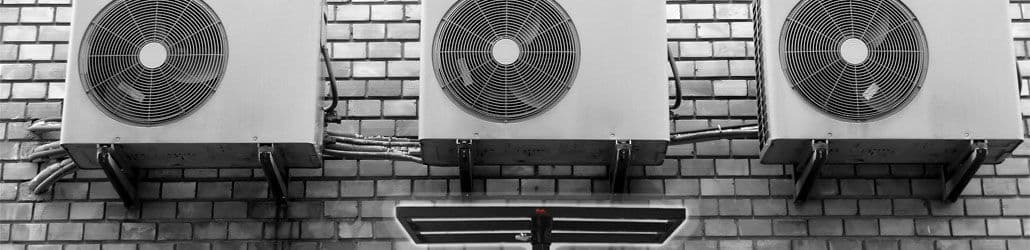 Drie airco-units gemonteerd aan een bakstenen muur, met leidingen zichtbaar en een materiaallift op de voorgrond.