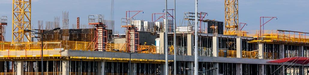 Grote bouwplaats met betonnen constructie, steigerwerk, bouwstempels en gele kranen op de achtergrond, typerend voor hoogbouw in de ruwbouwfase
