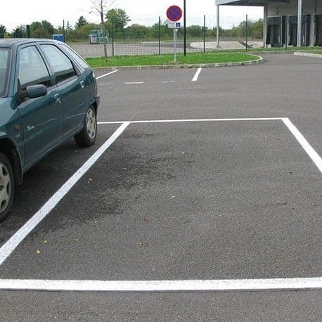 Witte lijnen markeren een parkeerplek op een geasfalteerd terrein, met een geparkeerde auto links en een verbodsbord in de achtergrond.