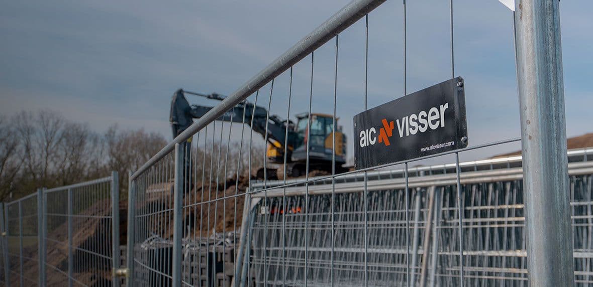 Metalen bouwhek met bedrijfsbord van AIC Visser voor een graafmachine op een zandige bouwplaats.