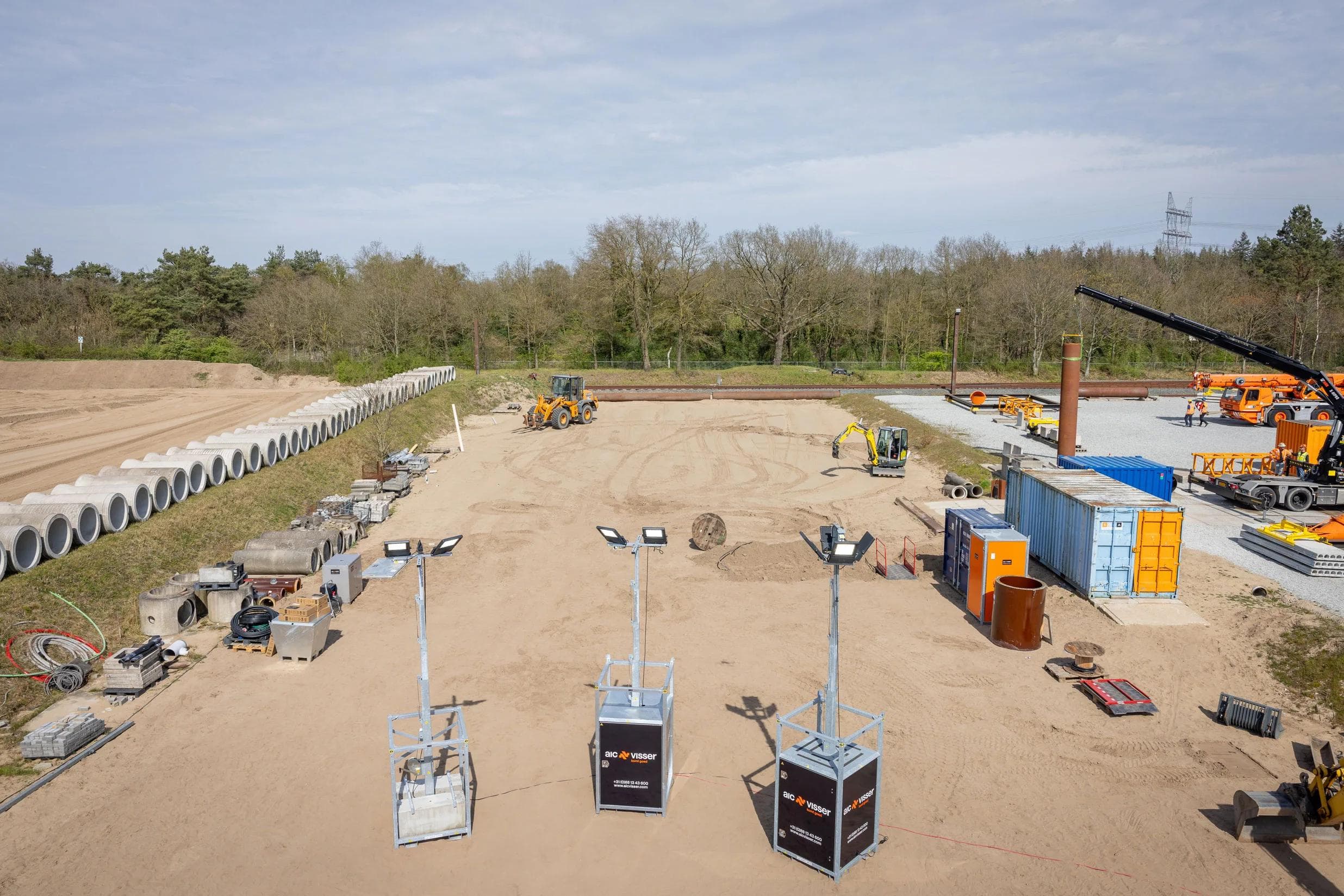 Bouwplaats met graafmachines, containers en gestapelde betonnen buizen op zandterrein.