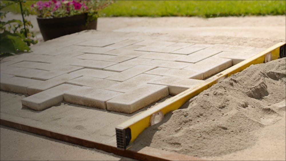 Grijze bestratingstegels worden gelegd op zand met een gele waterpas ernaast.